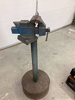 Full view of portable blue bench vise mounted on cylindrical metal stand with a wooden round base.