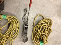 Top view of ratcheting come-along laying next to two yellow poly ropes coiled on floor, showing hooks and cable mechanism