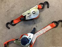 Close-up photo showing metal ratchet mechanism with orange webbing strap and black hooks on both ends.