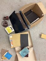 Multiple sandpaper sheets, emery cloth sheets, a blue abrasive pad, and rolls of abrasive material laid out on the floor showing various sizes and conditions.