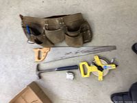 Tool belt and hand tools laid out on concrete floor showing belt, hammer, saw, tape measures, and measuring tools.