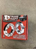 Box of Trans-Dapt billet aluminum wheel spacers and adaptors on concrete floor showing front with brand logo and product picture.