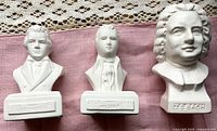 Three porcelain composer bust figures (Mozart, Beethoven, Bach) placed on a pink cloth with lace background, showing front and side details.