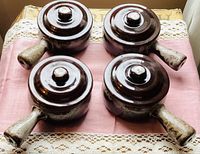 Top view of the four brown glazed pottery French onion soup bowls with lids and side handles arranged on a pink cloth with lace edges.