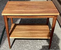 View of the teak two-tier side table showing the overall structure, warm wood tones, and condition of legs and shelves.