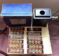 Complete set showing the antique metal magic lantern, 12 glass slides arranged in two rows, and original blue box.