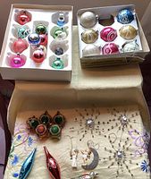 Two boxes of assorted glass baubles, including a blue bell, multiple circular baubles in various colours, and a handful of more uniquely shaped ornaments including stars and angels on a fabric surface.