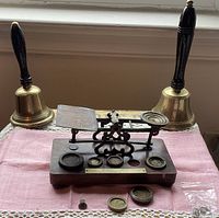 View of two brass bells with wooden handles flanking an antique postal scale on a wooden base with several brass weights placed on and in front of the base. The scale's brass measuring arm is detached from the wooden base.
