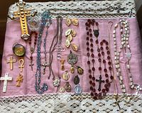 Top-down view of various religious crosses, medals, and rosaries displayed on pink fabric with lace edge. Items include large detailed crosses, crystal bead rosary, multiple medals and pendants.