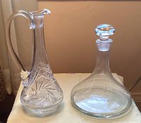 Two glass items on display: a clear glass decanter with swirl pattern, stopper, wide base, and a clear cut glass lead crystal pitcher with star patterns and curved handle.