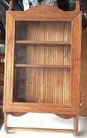 Closed front view of solid wood hanging display cabinet with glass door revealing three shelves and wood backing