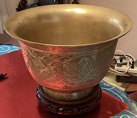 Large Asian brass and copper bowl with wide rim, showing engraved dragon design on side, sitting on carved wooden stand