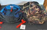 Two backpacks side by side on black surface, one blue and black with red straps, the other camouflage High Sierra backpack