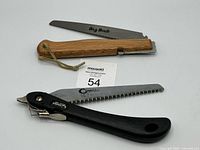 Two folding saws shown side by side on a white surface. One saw has a wooden handle with a leather strap, and the blade is marked Big Buck. The other saw has a black plastic handle and blade marked Coughlin 18-5.