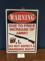 Front of the metal warning sign showing the printed caution message, shotgun image, and red and black text.