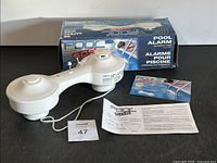 White SOS Pool Alarm device with attached cord placed near its original box and instruction manual on white surface.