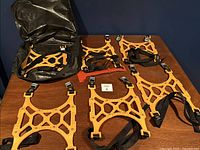 Full set of six orange rubber anti-slip traction mats with metal studs laid out on a wooden table, showing black adjustable straps and metal buckles, and a black storage bag in background.