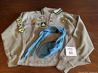 Light brown Wolf Cubs uniform shirt laid flat showing embroidered patches on chest and sleeves, blue neck scarf folded on the shirt.