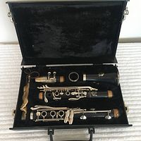 Open black hard case showing disassembled clarinet body sections (upper joint, lower joint, bell) with silver keys and cork joints inside plush black interior lining.