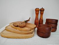 Photo showing stacked three rectangular wood serving boards, wooden salad servers, five small stacked wood bowls, salt shaker, and pepper grinder arranged on white cloth.