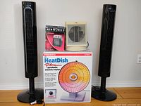Five electric devices including two tall black Honeywell tower fans, Presto HeatDish Plus parabolic heater in box, small cream Airworks ceramic heater, and Honeywell thermostatic heater.