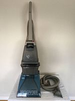 Hoover Steam Vac upright carpet cleaner in blue and gray with clear dirt chamber shown standing upright with hose and tool attached at base.