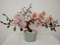 Full view of the ornamental Chinese glass jade bonsai tree with pink, peach, white flowers and green leaves in a hexagonal pale green ceramic pot with white sand.