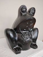 Front view of black cement frog-shaped garden planter showing sculpted eyes, feet, and floral relief on belly.