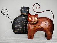 Front view of two hollow metal cat sculptures showing black and copper finishes with curled tails and perforated bodies.
