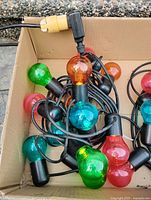 Close-up image of multi-colored bulbs including green, orange, red, and blue on black string light with black sockets, showing electrical plug.