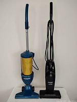 Front full view of two Eureka corded bagless stick vacuums, one blue and one black, standing upright on a white surface.