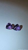Pair of matched natural Brazilian amethyst gemstones, trillion cut, placed side by side on a plain surface, showing the deep purple color and clarity.