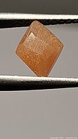 Diamond-shaped peach moonstone held by tweezers showing surface and color