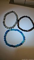 Three natural gemstone stretchy bracelets arranged on a flat surface, showing their round beads in blue, black/brown, and gray-green hues.