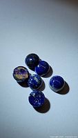 Six round natural lapis lazuli cabochon stones with distinctive deep blue color and flecks of pyrite, arranged loosely on a white surface.