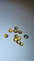 Natural small round cabochon-cut rainbow sapphires in yellow, orange, and green shades, displayed on a light surface under light. The stones vary slightly in size, about 2.7mm in diameter.