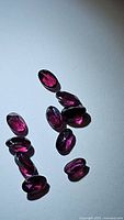 Lot of 11 natural oval cut rhodolite garnet gemstones, purple-pink color, shown on a white background.