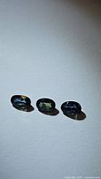 Photo shows three oval cut sapphires in blue and green hues laid out on a white background.