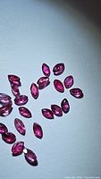 Close-up photo of 24 small marquis-cut rubies spread on a white surface, showing their deep pink-red hues and faceted details.
