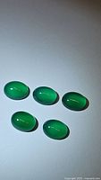 Five oval vivid green onyx cabochon gemstones evenly spaced on white background with light reflection showing smooth polish and clarity.