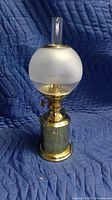 Full front view of antique brass oil lamp on quilted blue blanket, showing frosted glass globe and clear glass chimney.