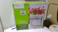 Boxed new Flower Box Plant Stand with green and white packaging, showing a black metal frame with wire basket top filled with flowers on the box image.