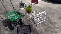 Photo showing garden spreader, several metal bird houses and feeders, ceramic bird feeder, white metal plant stands and a black tray on concrete surface.