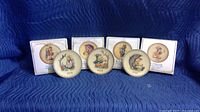 Seven Hummel collectable plates arranged on a blue quilted surface. Four plates are in front of their original white boxes with illustrations and text. Plates have dated motifs and classic Hummel designs.