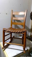 Photo of a small wooden rocking chair for kids with ladder back and slatted seat, showing front view on a white surfaced table against a plain wall.