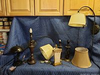 All seven lamps and lamp shades grouped together on a blue quilted blanket; shows variety of styles and materials including metal, ceramic, and fabric shades