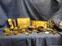 Wide shot showing entire metal collection with lion figurine, paintings, trays, duck sculpture, match holders, belt buckle, and other metal items arranged on a blue quilted blanket.