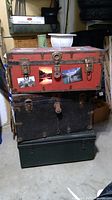 Side view of the three stacked trunks showing the red steamer trunk on top with stickers and locks, the black trunk in the middle, and the green metal foot locker at the bottom.
