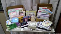 Photo showing a large collection of greeting cards, stationery, notepads, book covers, address book, and inspirational books arranged on a table with two open boxes.