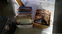 Two cardboard boxes on floor, one filled with approximately 50 vintage 78 RPM records in brown paper sleeves, other with about 50 assorted 45 RPM and other records